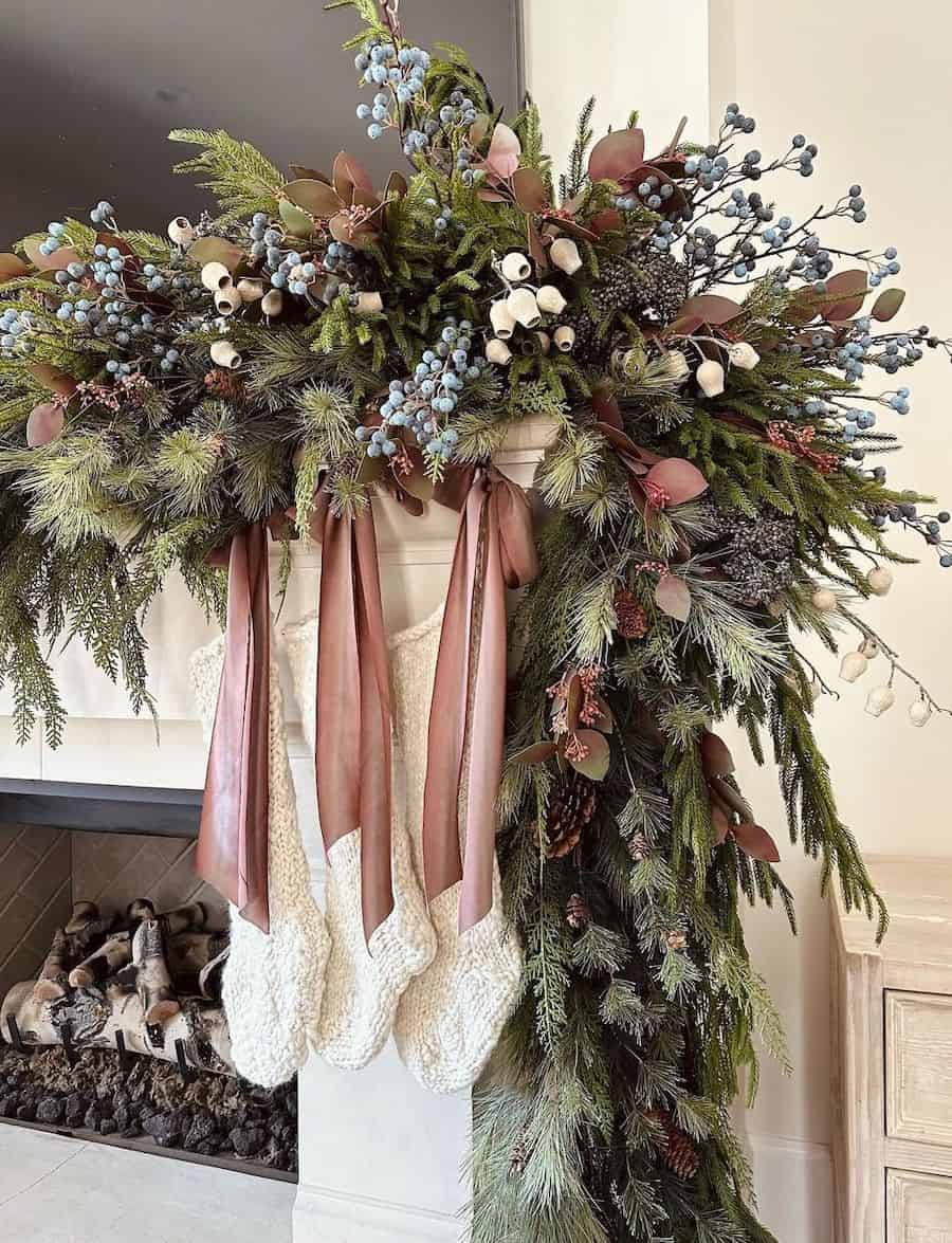a christmas mantle with pine garland featuring pinecones, pink ribbons, white flowers, and blueberries