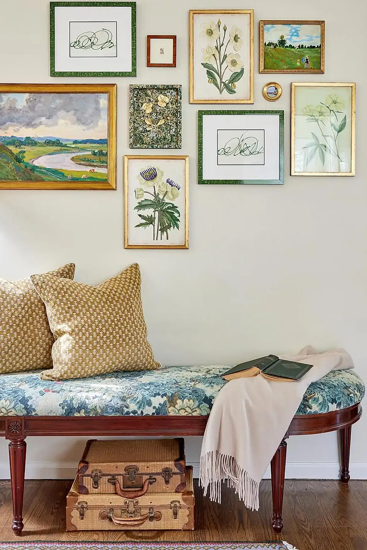 Entryway with floral-upholstered bench, gold and green framed art, patterned pillows, and vintage suitcases underneath.