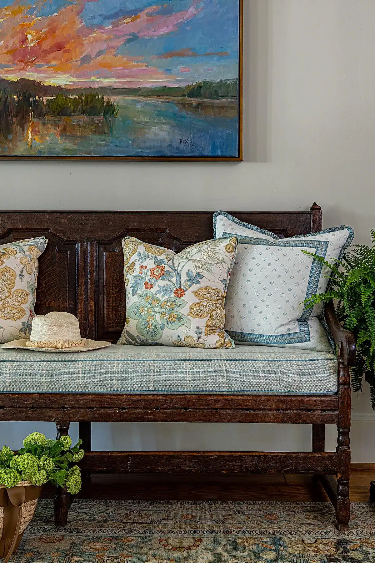 Antique wooden bench with floral-patterned pillows beneath a colorful landscape painting in an entryway.