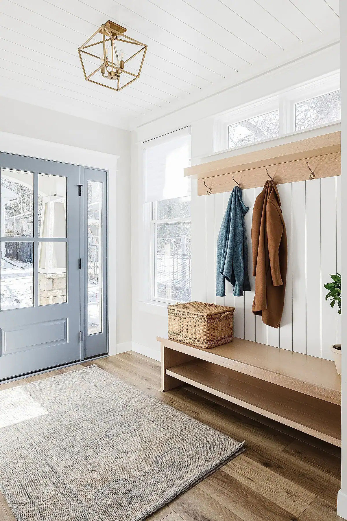 Entryway with wooden bench, wicker basket, light-colored rug, and hooks on white paneling.