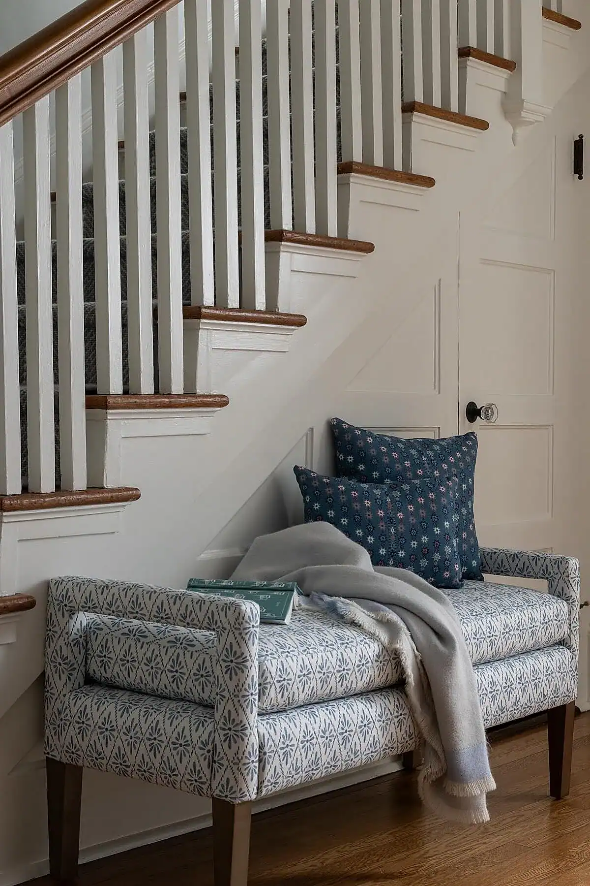 Upholstered bench with blue patterned fabric, decorative pillows, and blanket under a staircase in an entryway.