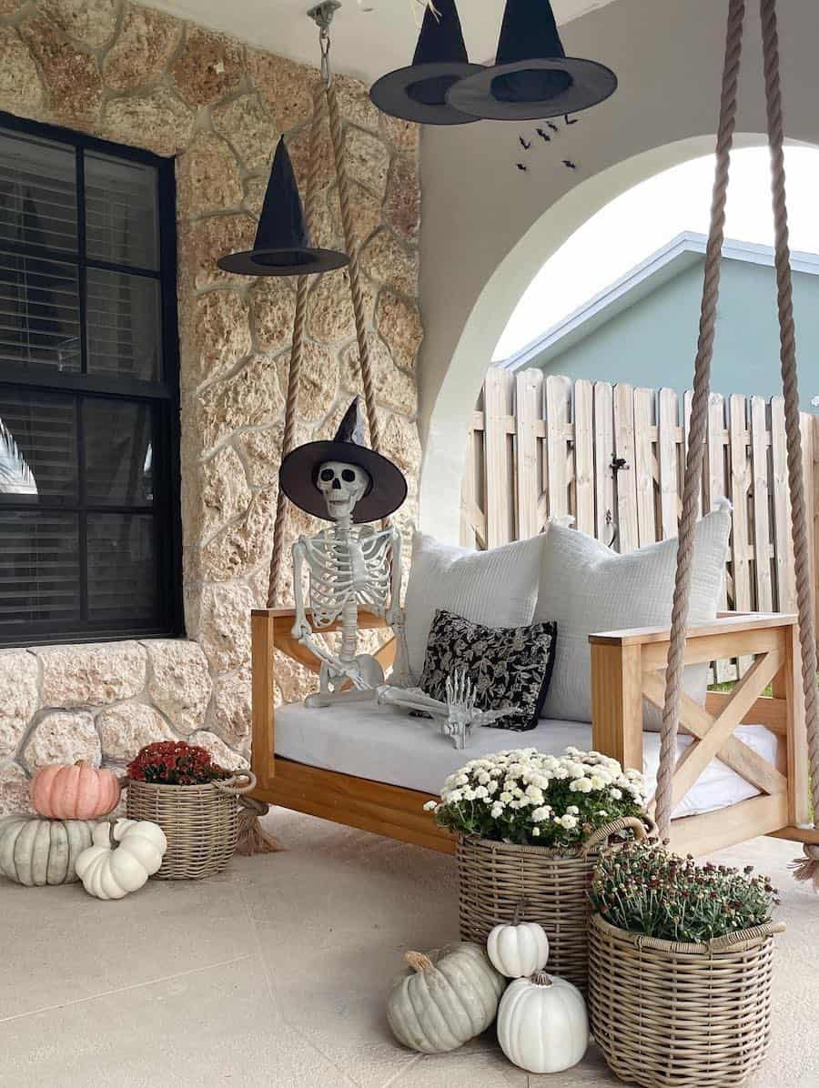 a front porch featuring a swing with a skeleton wearing a witch hat sitting on it, pumpkins, florals, and more floating witch hats
