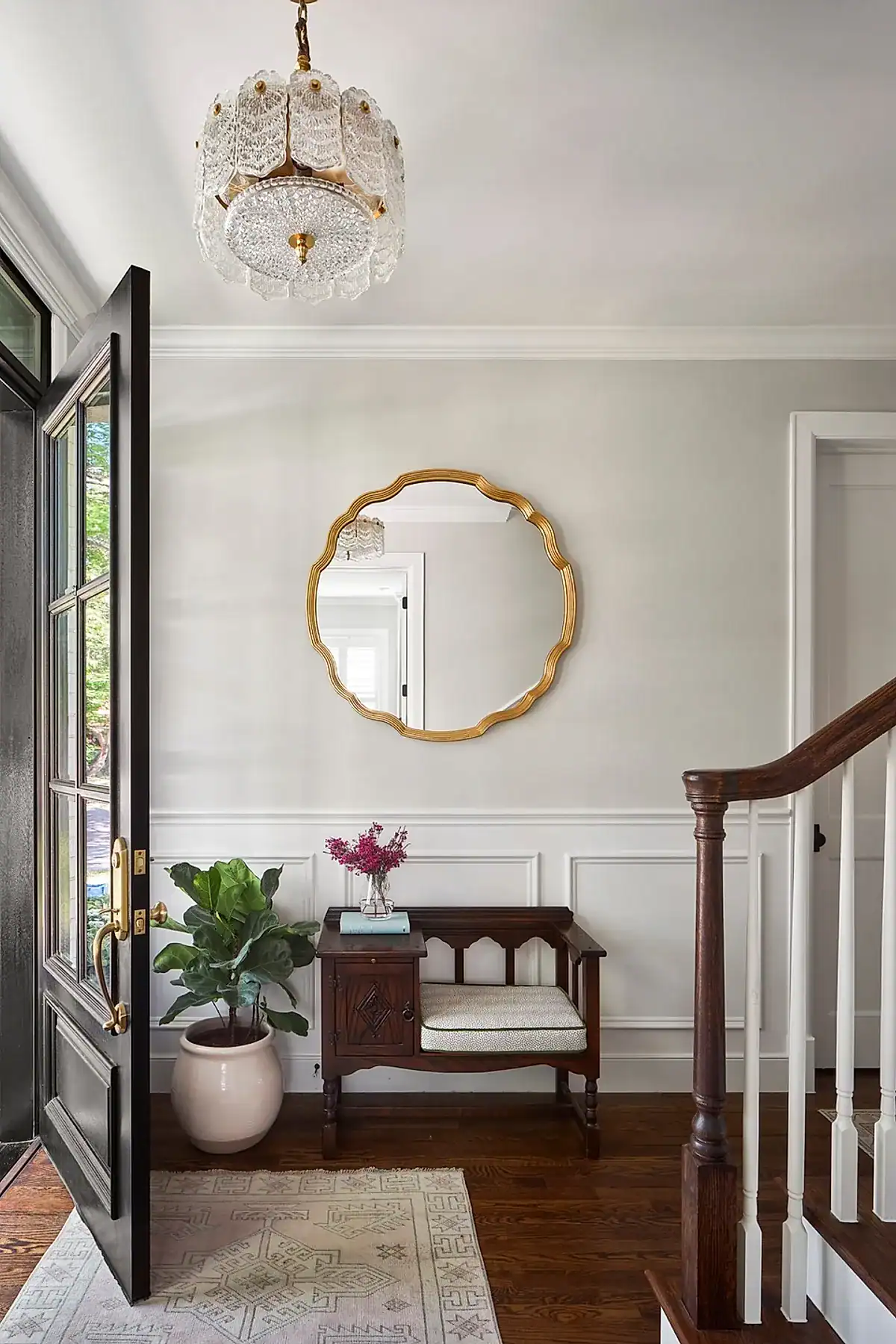 Entryway with a wooden bench and cushion, gold mirror, crystal chandelier, and potted plant next to an open door.